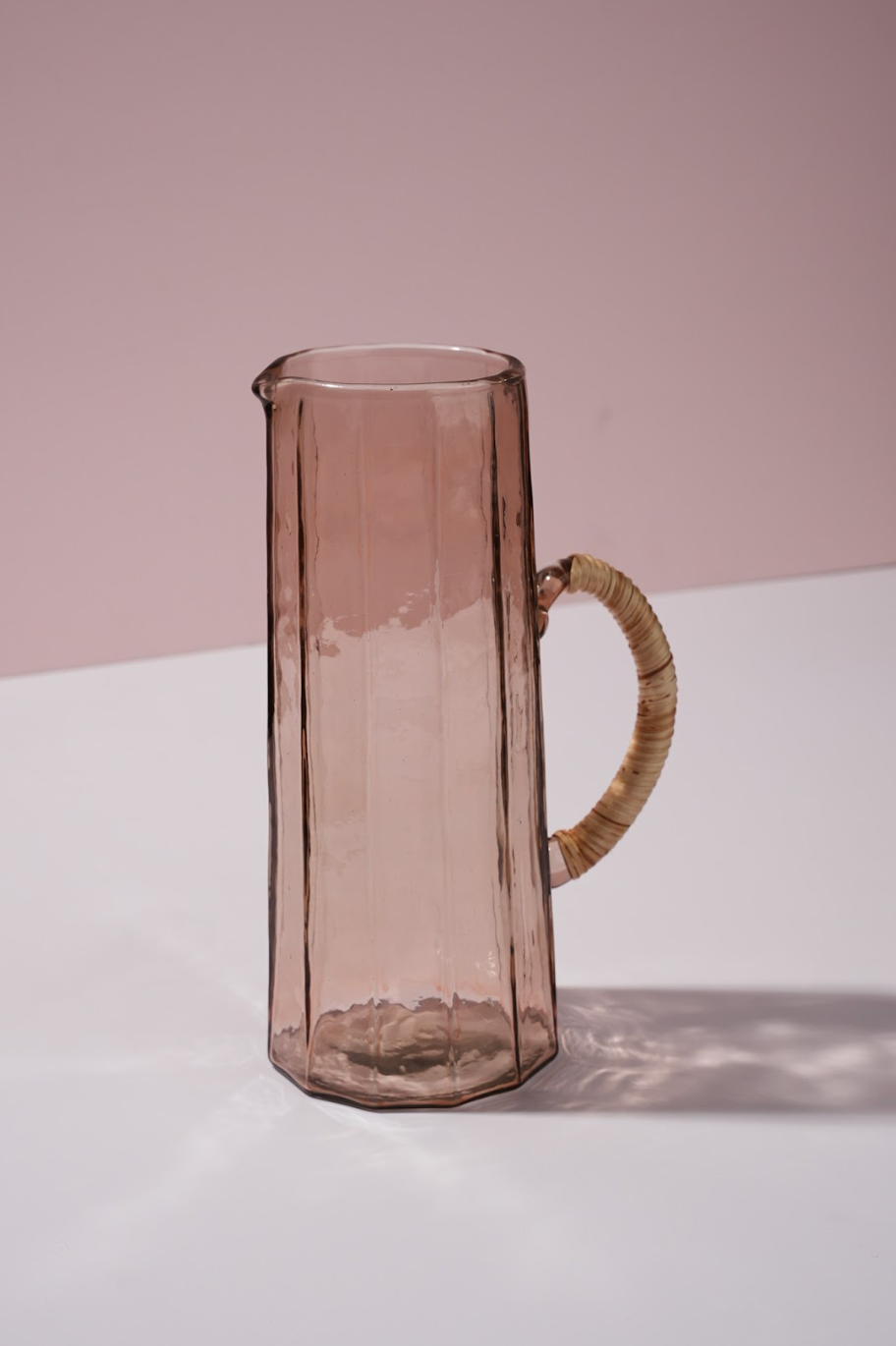 Pink glass pitcher with wrapped bambo handle on a white surface and pink background